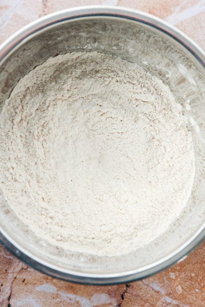 A metal mixing bowl filled with flour sits on a brown countertop. The flour appears to be evenly distributed and slightly textured.