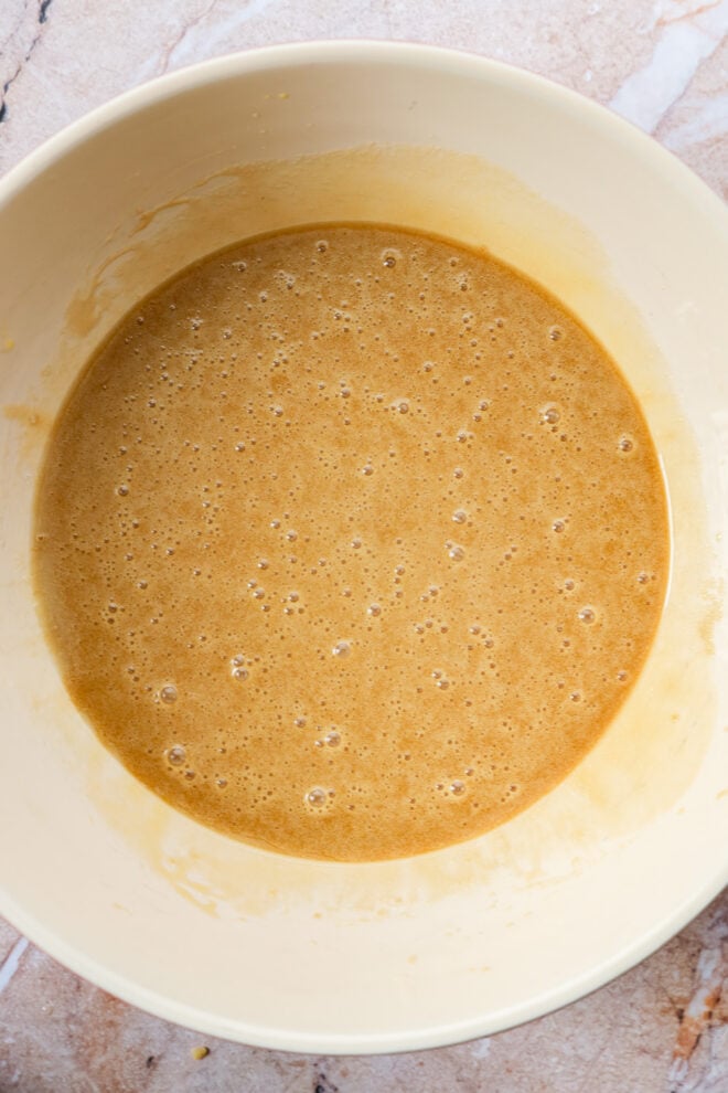 A creamy, light brown carrot cake loaf batter with small bubbles sits in a large, off-white mixing bowl on a marble countertop.