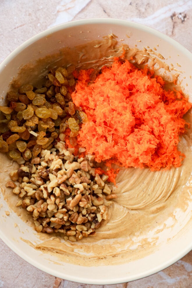 A mixing bowl with partially mixed carrot cake loaf batter, topped with shredded carrots, chopped walnuts, and golden raisins, on a light marble surface.