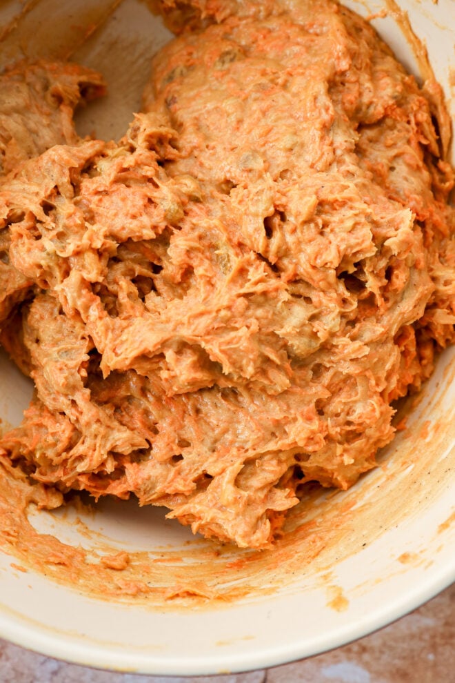 A close-up of a mixing bowl filled with thick, orange-brown batter, with visible shreds of carrot, suggesting a mixture for a delicious carrot cake loaf or muffins.
