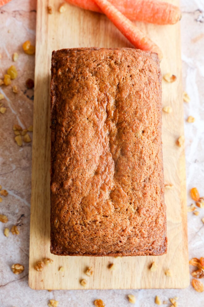A carrot cake loaf rests on a wooden cutting board, surrounded by two whole carrots, raisins, and chopped walnuts scattered across a marble surface.