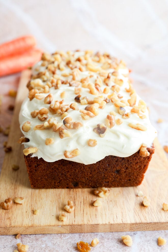 A carrot cake loaf with cream cheese frosting and chopped walnuts on top sits on a wooden cutting board. Two carrots are blurred in the background.