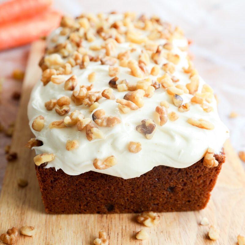 A carrot cake loaf with cream cheese frosting and chopped walnuts on top sits on a wooden cutting board. Two carrots are blurred in the background.