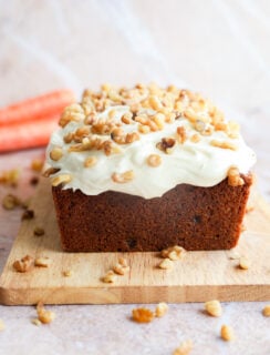 A carrot cake loaf with cream cheese frosting and chopped walnuts sits on a wooden cutting board, with fresh carrots visible in the background.