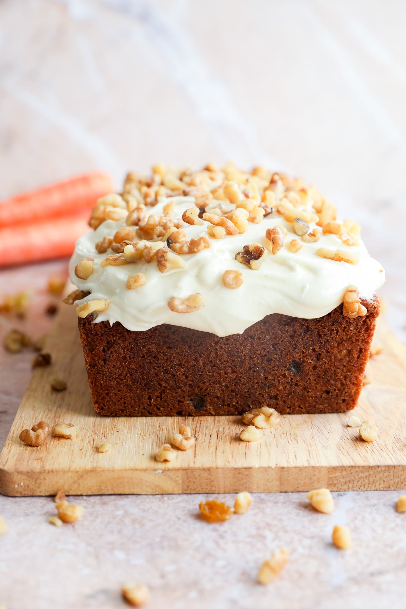 A carrot cake loaf with cream cheese frosting and chopped walnuts sits on a wooden cutting board, with fresh carrots visible in the background.