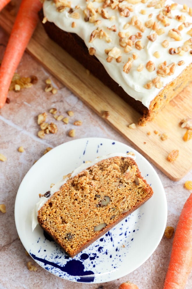 A slice of carrot cake loaf with cream cheese frosting and walnuts sits on a white plate with a blue splatter design. The rest of the cake is on a wooden board nearby, surrounded by whole carrots and scattered walnuts.