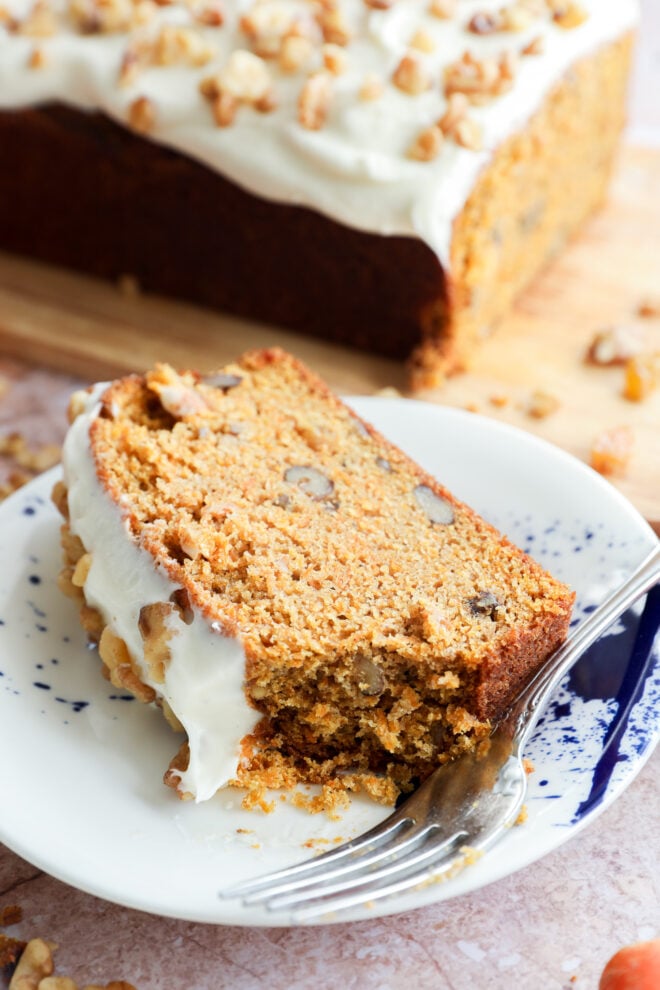 A slice of carrot cake loaf with cream cheese frosting and chopped walnuts sits on a white plate with a fork, while the rest of the loaf is in the background on a wooden board.
