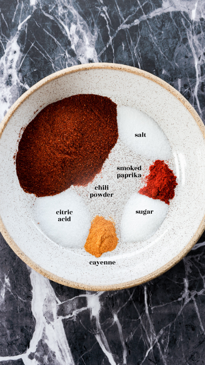 A white plate on a marble surface holds separate piles of chili powder, smoked paprika, cayenne, citric acid, salt, and sugar&mdash;each labeled with text&mdash;perfect ingredients for making homemade chili lime seasoning.