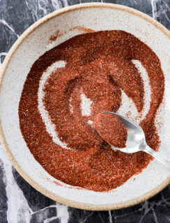 A spoon mixes a vibrant chili lime seasoning, a reddish-brown spice blend, in a round, white bowl set on a dark marble surface.