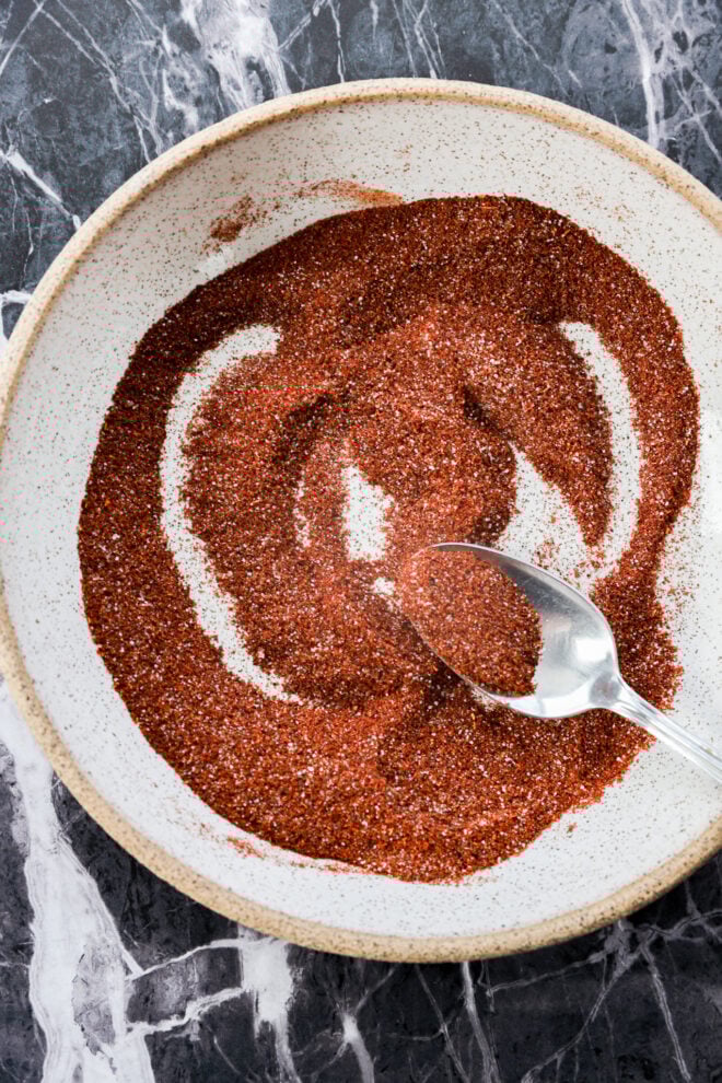 A spoon mixes a vibrant chili lime seasoning, a reddish-brown spice blend, in a round, white bowl set on a dark marble surface.