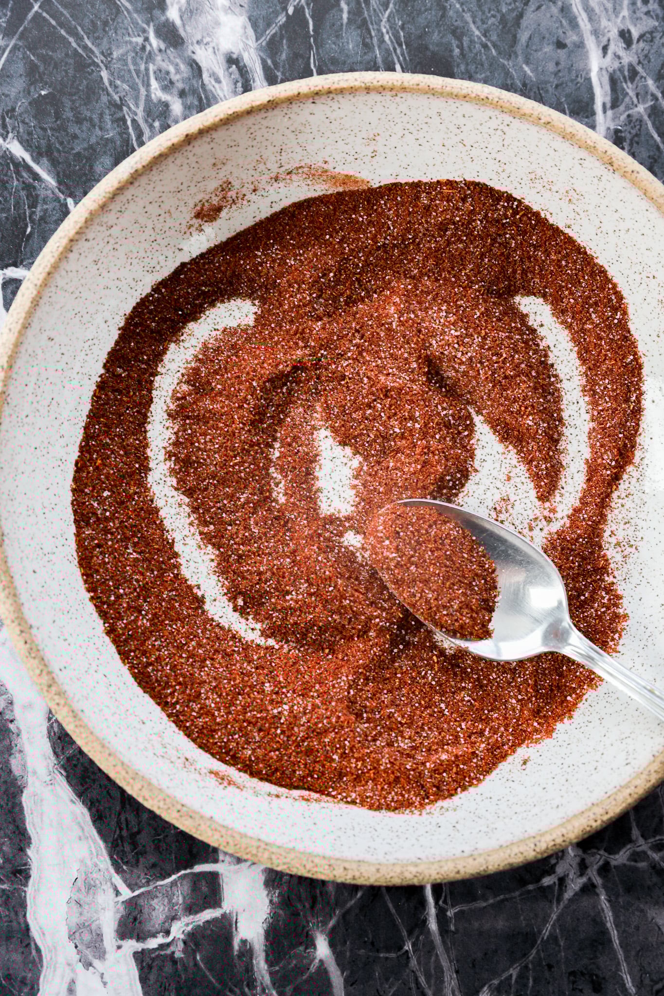 A spoon mixes a vibrant chili lime seasoning, a reddish-brown spice blend, in a round, white bowl set on a dark marble surface.