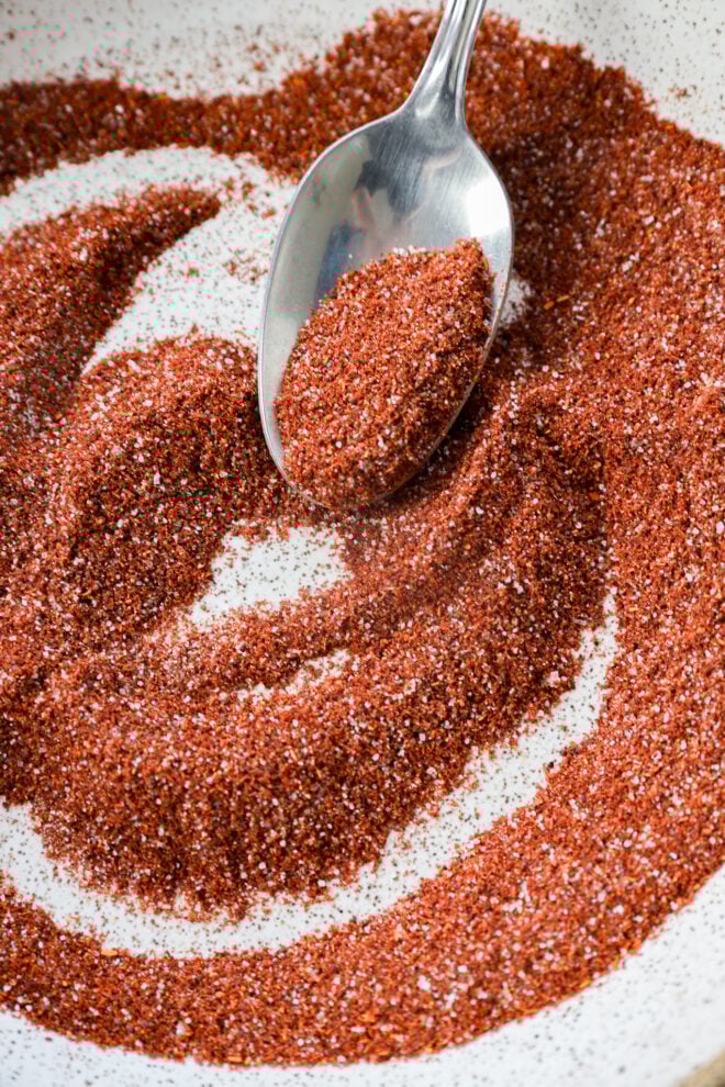 A close-up of a spoon scooping chili lime seasoning from a white bowl, with the reddish-brown mixture spread out in swirling patterns.