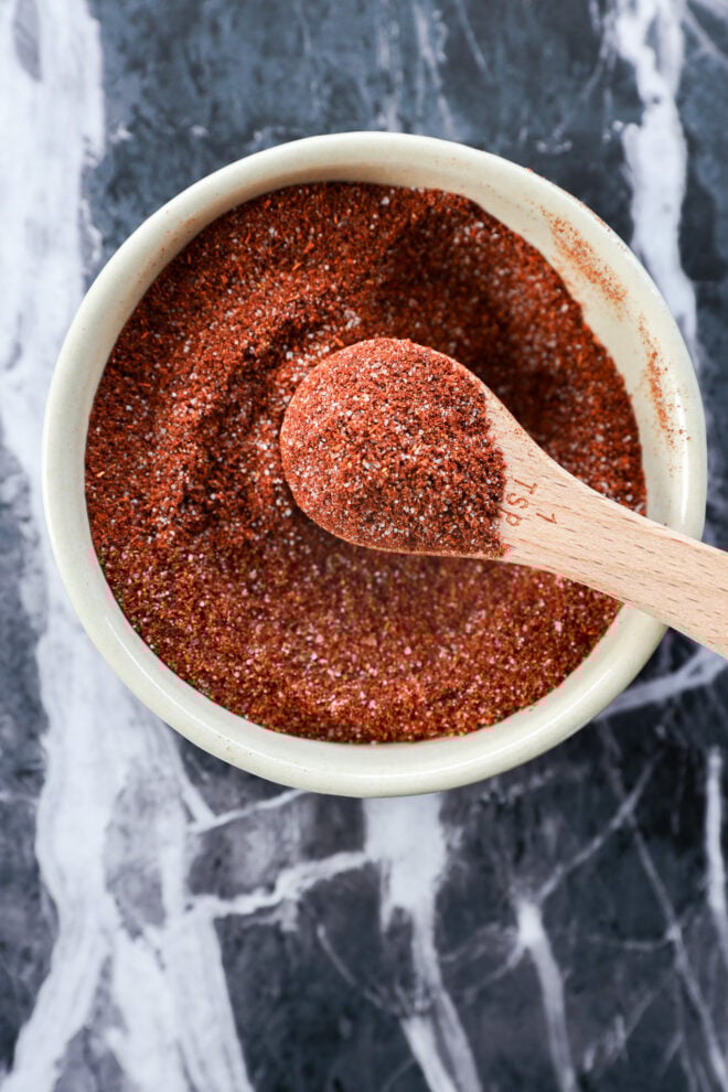 A white bowl filled with reddish-brown chili lime seasoning, with a wooden measuring spoon resting inside and holding some of the zesty spice mix, on a dark marbled surface.