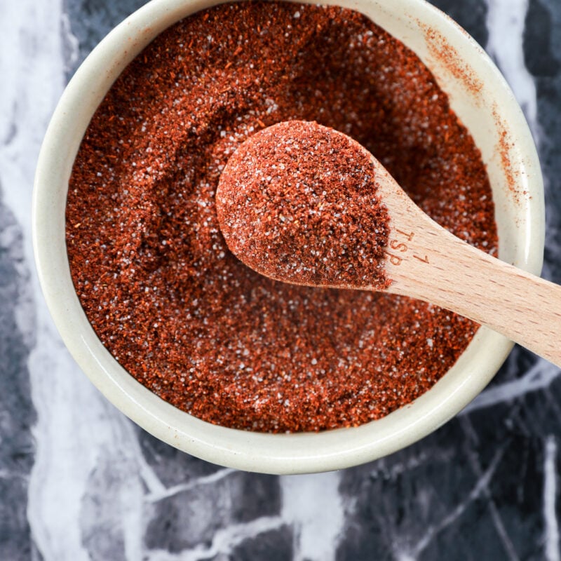 A white bowl filled with reddish-brown chili lime seasoning, with a wooden measuring spoon resting inside and holding some of the zesty spice mix, on a dark marbled surface.