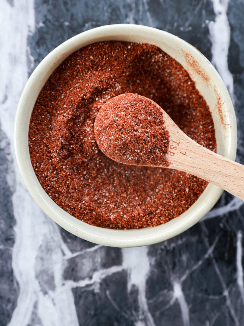 A white bowl filled with reddish-brown chili lime seasoning, with a wooden measuring spoon resting inside and holding some of the zesty spice mix, on a dark marbled surface.