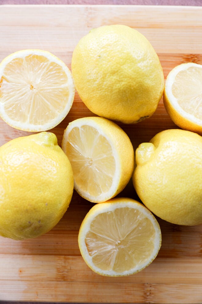 Six lemons, three whole and three cut in half, are arranged on a wooden cutting board.