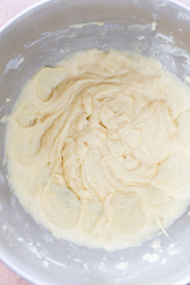 A metal mixing bowl filled with smooth, light yellow lemon loaf cake batter that has been partially mixed, with swirls and ridges visible on the surface.
