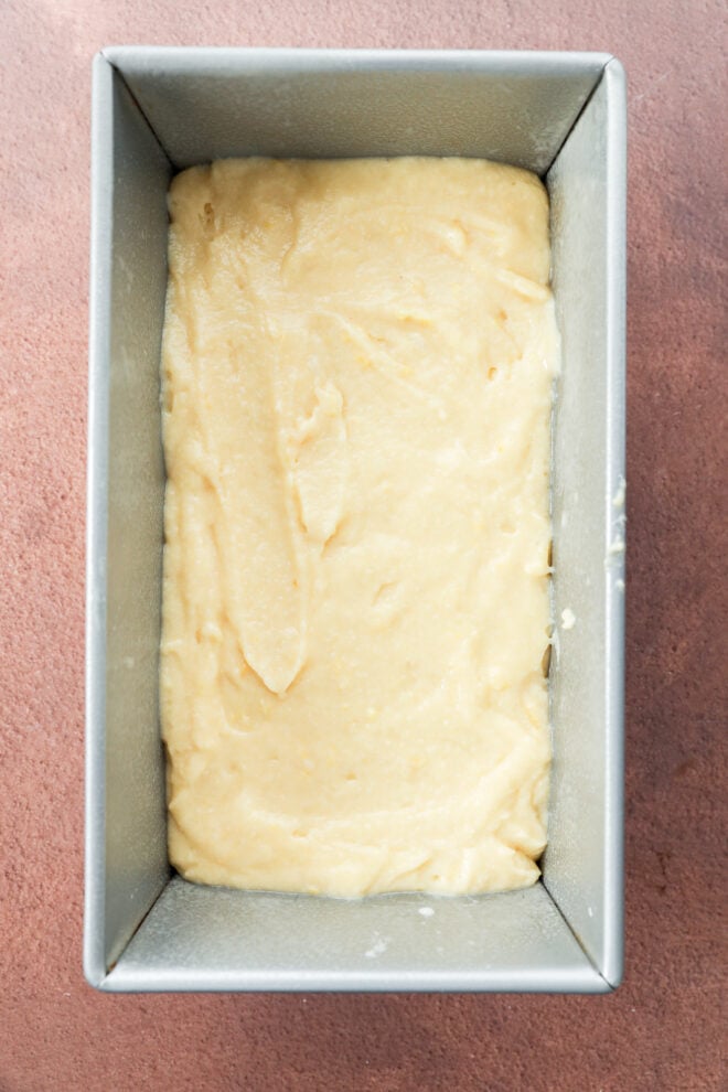 A metal loaf pan filled with smooth, pale yellow batter, ready to be baked, sits on a brown textured surface.