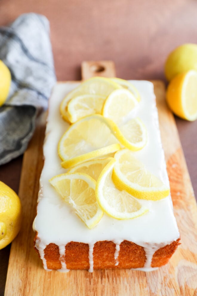 A delicious lemon loaf cake topped with white icing and lemon slices sits on a wooden board, surrounded by fresh lemons and a gray cloth in the background.