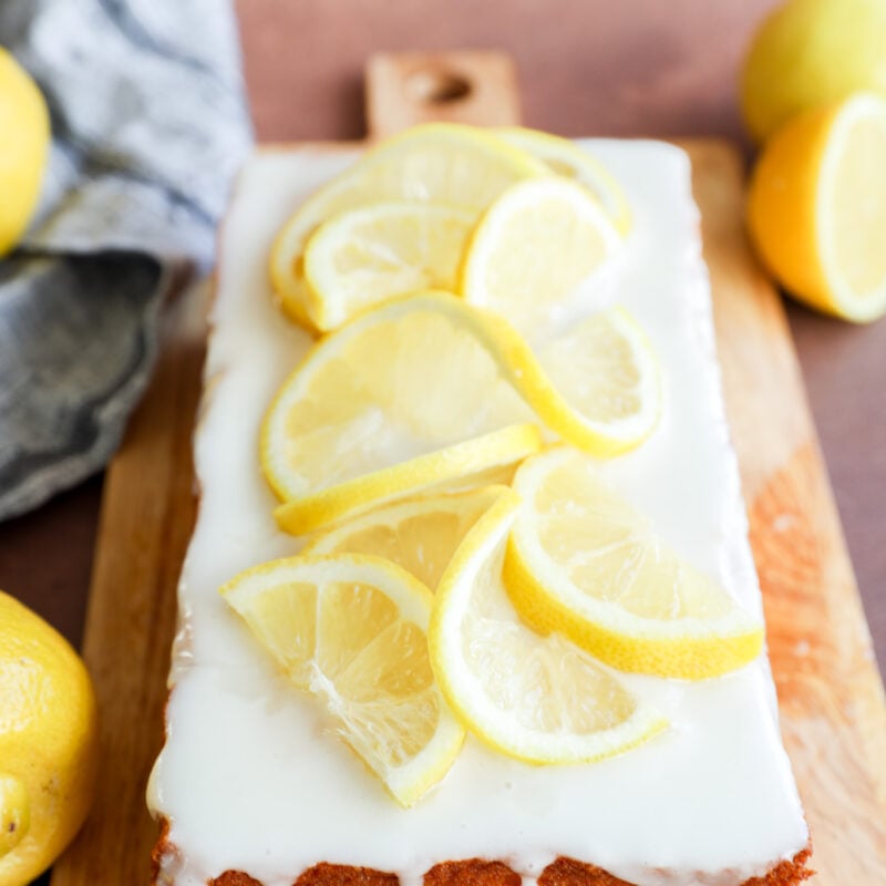 A delicious lemon loaf cake topped with white icing and lemon slices sits on a wooden board, surrounded by fresh lemons and a gray cloth in the background.