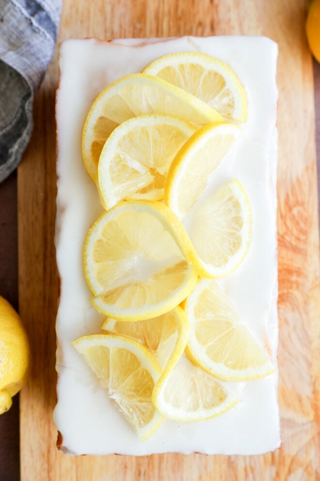 A rectangular dessert with white icing, topped with overlapping slices of fresh lemon, sits on a wooden board. A whole lemon and a gray-striped cloth are partially visible nearby.