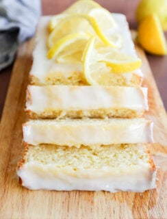 A lemon loaf cake with white glaze, sliced and topped with fresh lemon slices, is displayed on a wooden cutting board. Lemons are visible in the background.