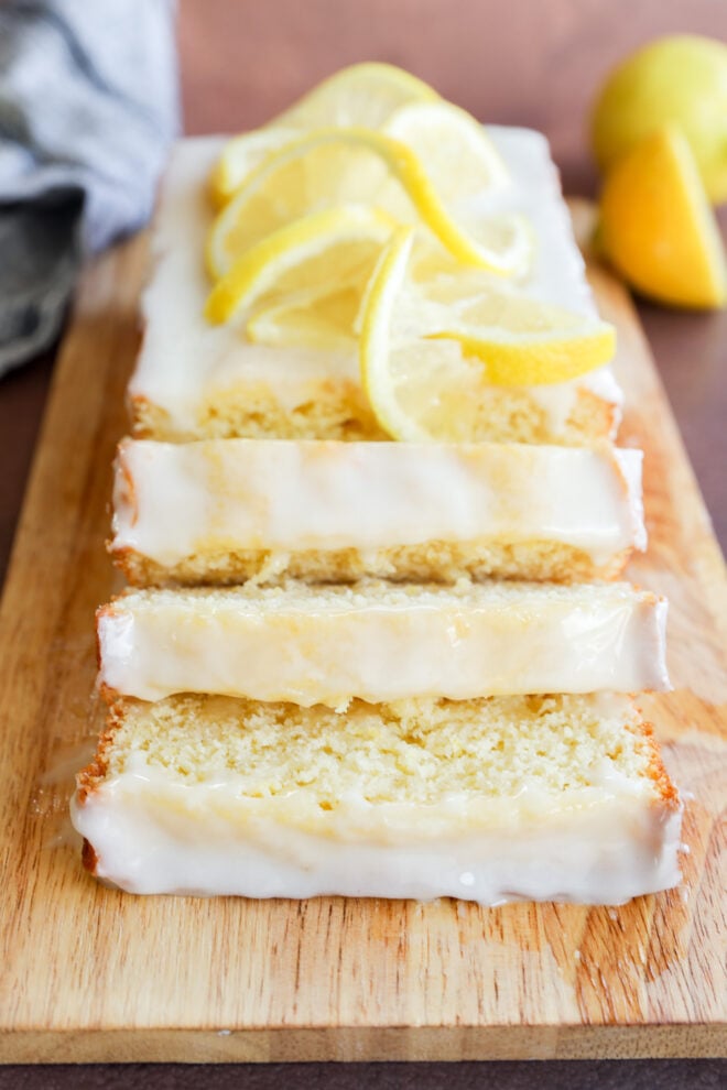 A lemon loaf cake with white glaze, sliced and topped with fresh lemon slices, is displayed on a wooden cutting board. Lemons are visible in the background.
