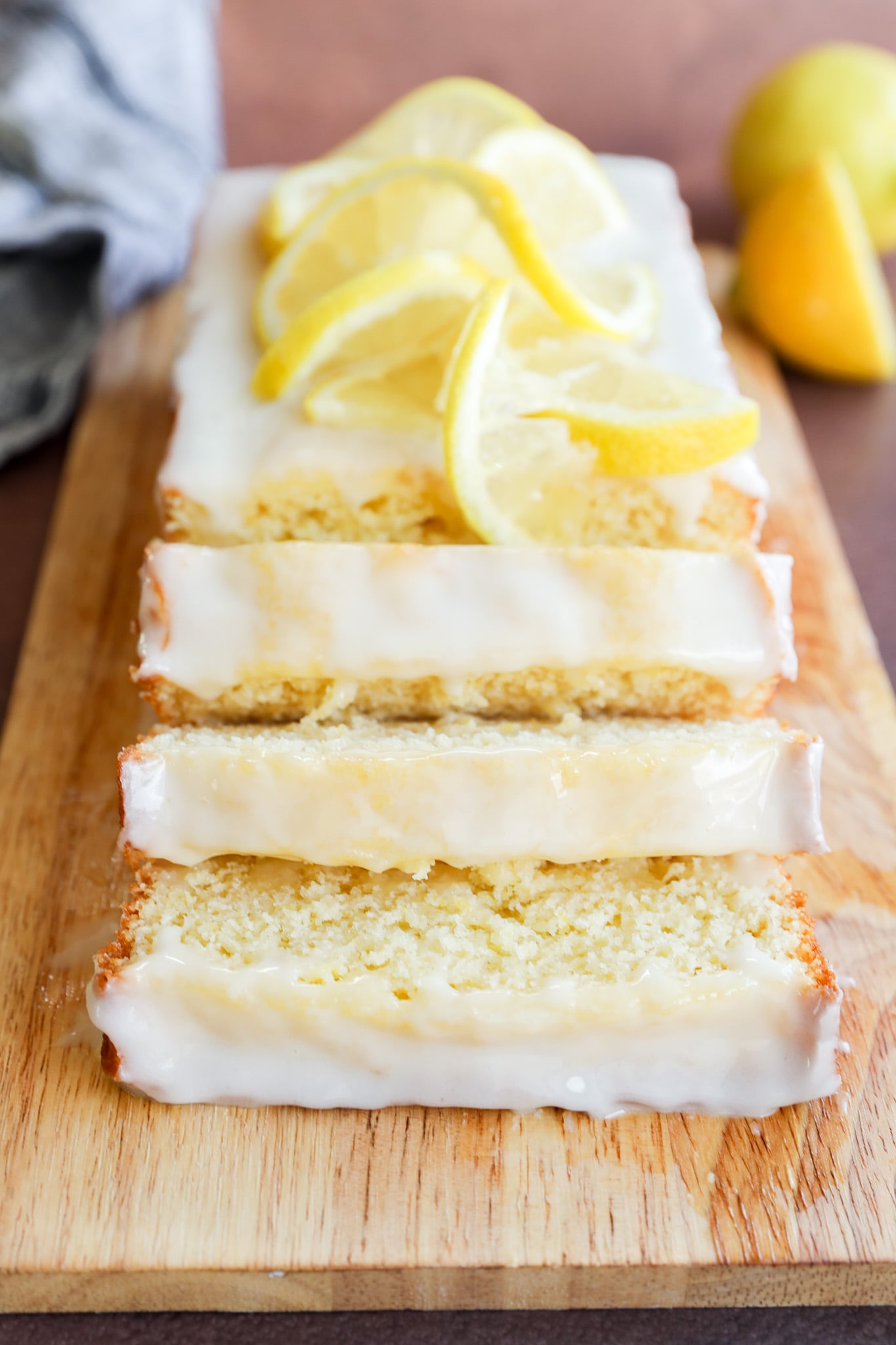A lemon loaf cake with white glaze, sliced and topped with fresh lemon slices, is displayed on a wooden cutting board. Lemons are visible in the background.