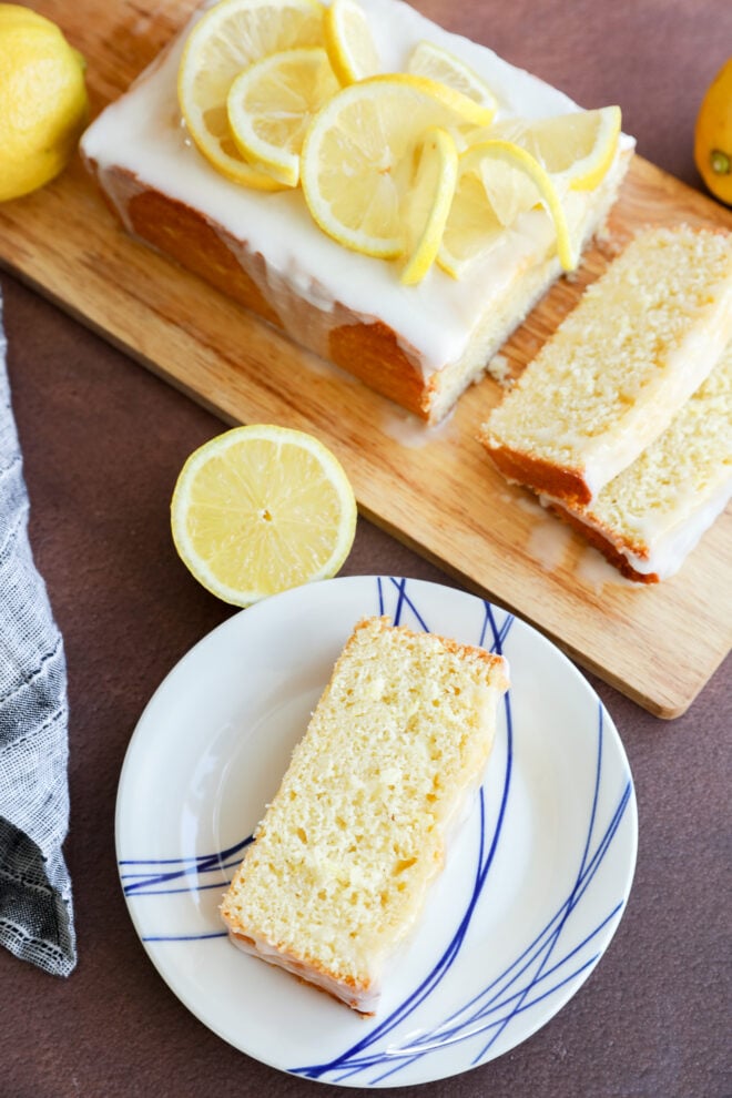 A lemon loaf cake with white icing and lemon slices on top sits on a wooden board, while a partially sliced piece rests on a white plate with blue lines and a halved lemon nearby.