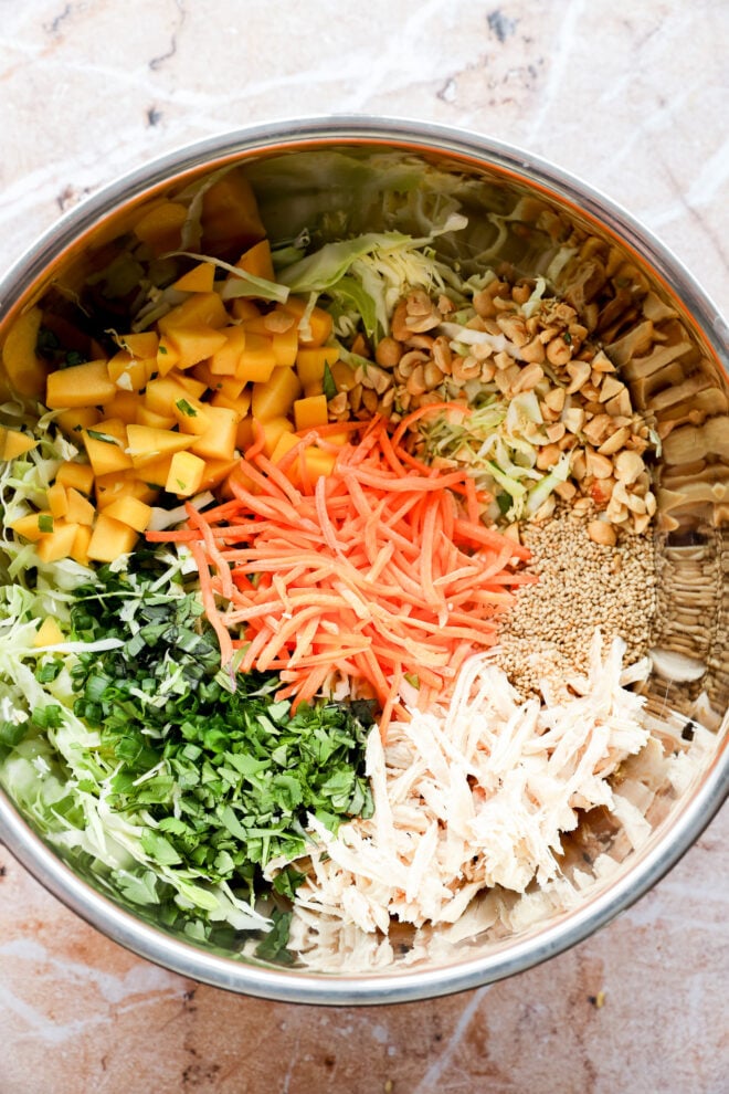 A large metal bowl filled with shredded cabbage, diced mango, chopped peanuts, shredded carrots, sesame seeds, chopped cilantro, and shredded chicken, arranged in separate sections.