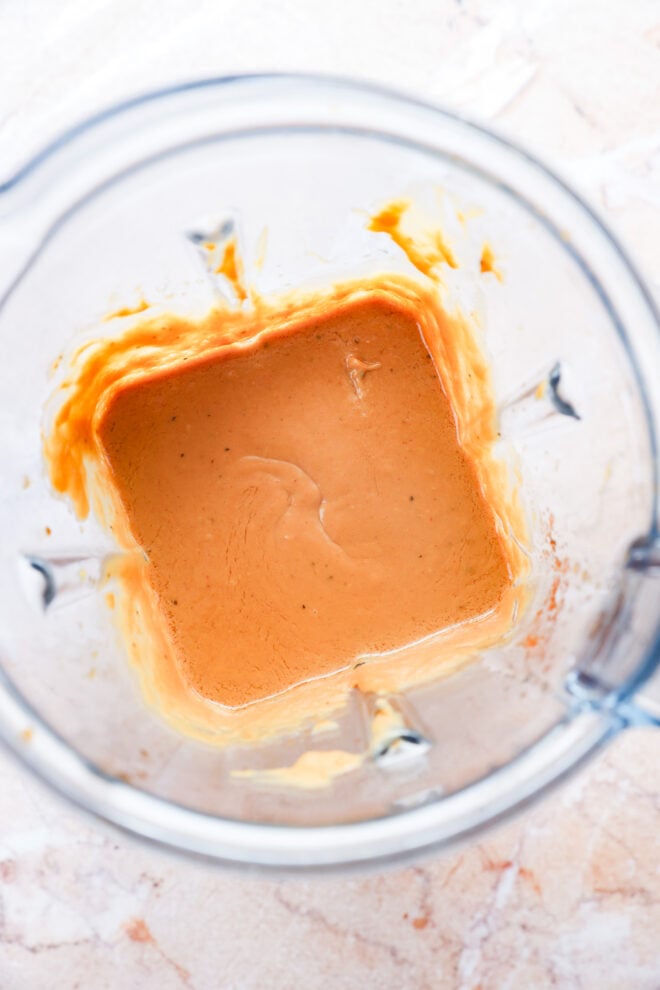 Top-down view of a blender containing a creamy, orange-colored sauce or dressing, with some sauce splattered along the sides of the clear blender container.