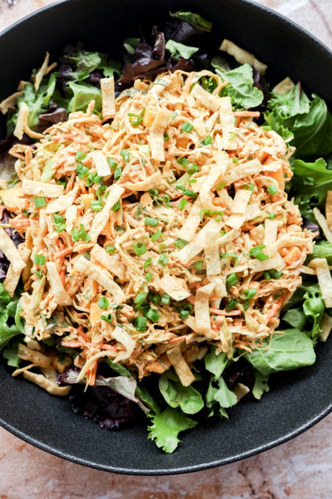 A black bowl filled with mixed greens topped with shredded chicken, thin crispy tortilla strips, grated carrots, and a sprinkling of chopped green onions.