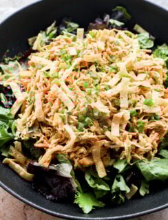 A black bowl filled with mixed greens, shredded chicken, julienned carrots, crispy tortilla strips, and topped with chopped green onions.