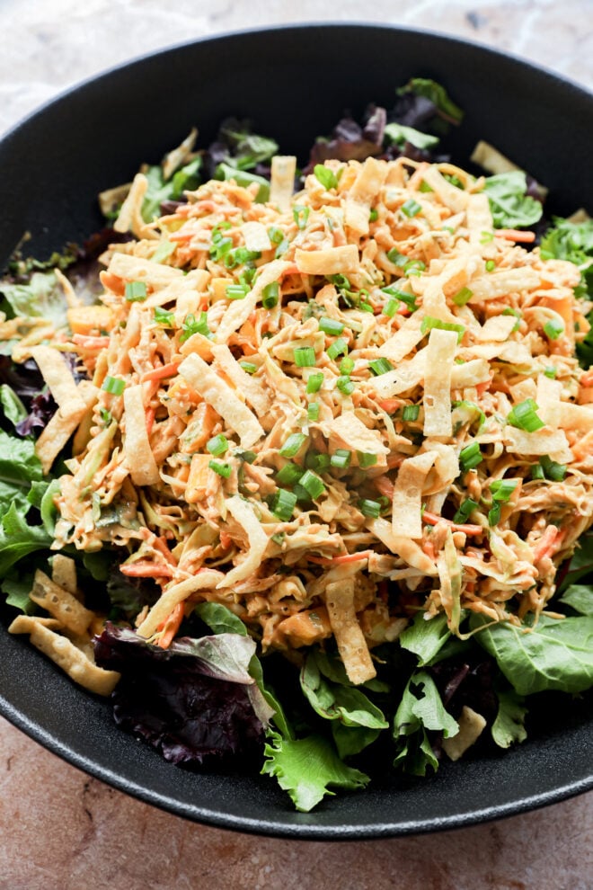A black bowl filled with mixed greens, shredded chicken, julienned carrots, crispy tortilla strips, and topped with chopped green onions.