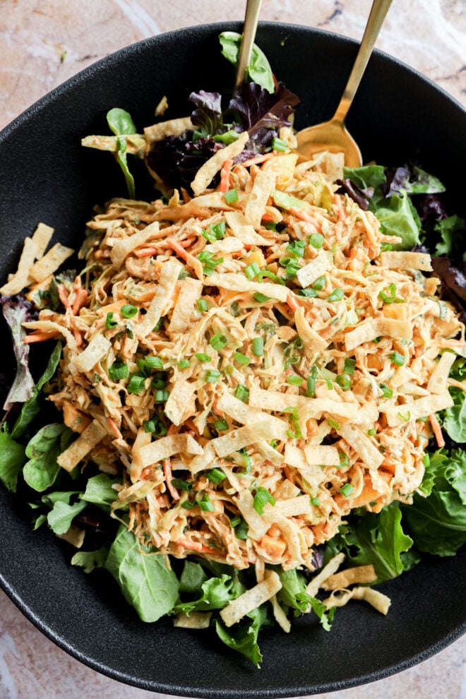 A black bowl filled with mixed greens topped with shredded chicken, crispy tortilla strips, and chopped chives, with two gold utensils resting in the salad.