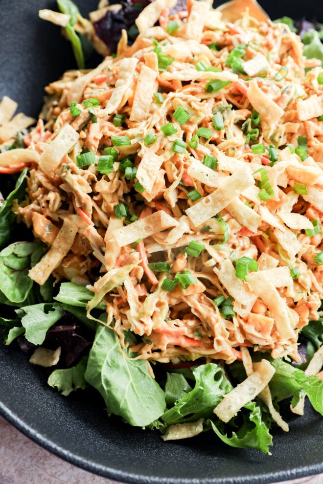A close-up of a salad topped with shredded chicken, tortilla strips, chopped green onions, and a creamy dressing, served on a bed of mixed leafy greens in a black bowl.