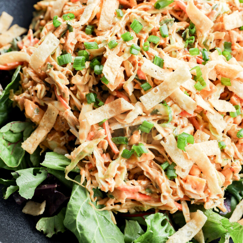 A close-up of a salad topped with shredded chicken, tortilla strips, chopped green onions, and a creamy dressing, served on a bed of mixed leafy greens in a black bowl.