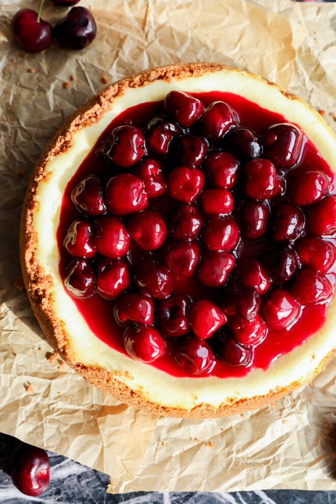 A whole summer dessert sits on crumpled parchment paper, topped with glossy cherries and fruit glaze. A few cherries are scattered nearby, adding to the tempting display of this classic dessert.