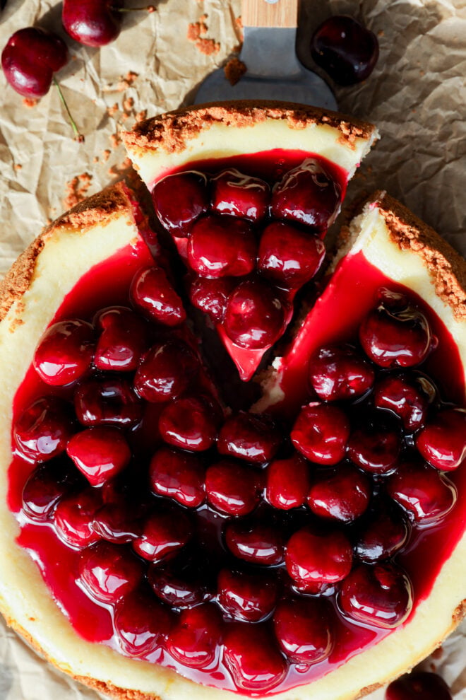 A classic Cherry Cheesecake with a graham cracker crust, topped with glossy, whole cherries in red syrup, sits on parchment paper. One slice of the cherry cheesecake is partially cut and slightly pulled apart from the whole cake.