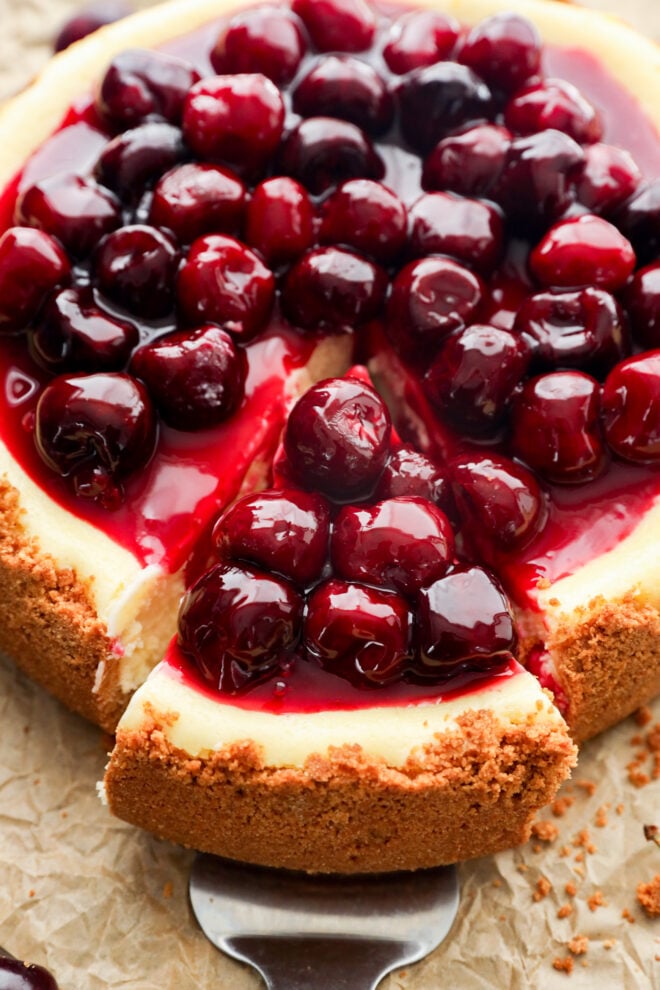 A summer dessert with a graham cracker crust is topped with glossy cherries and sauce; a slice has been cut and is being served, revealing the creamy interior.