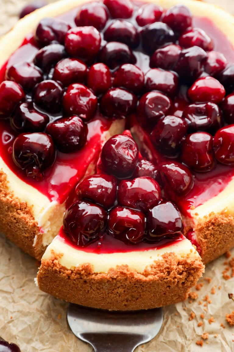 A summer dessert with a graham cracker crust is topped with glossy cherries and sauce; a slice has been cut and is being served, revealing the creamy interior.