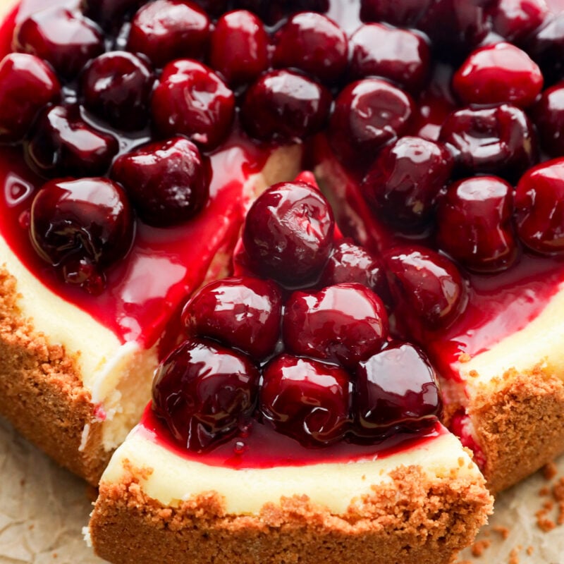 A summer dessert with a graham cracker crust is topped with glossy cherries and sauce; a slice has been cut and is being served, revealing the creamy interior.