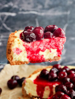 A close-up of Cherry Cheesecake with a graham cracker crust, creamy filling, cherry topping, and glossy red sauce, being lifted from the whole cake in the background.