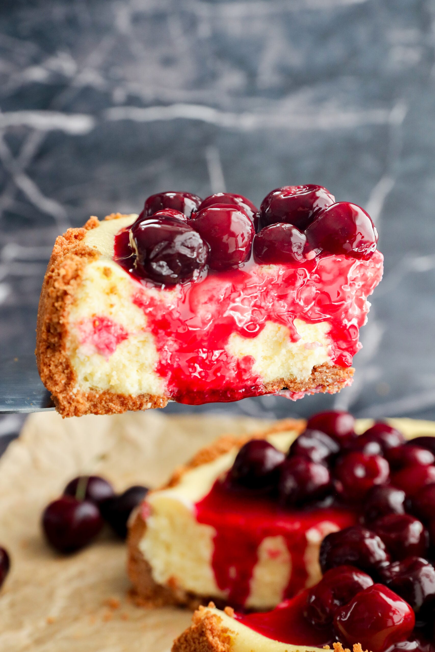 A close-up of Cherry Cheesecake with a graham cracker crust, creamy filling, cherry topping, and glossy red sauce, being lifted from the whole cake in the background.