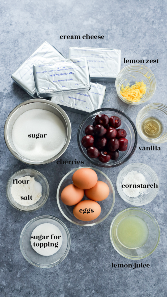 Ingredients for Cherry Cheesecake are arranged on a gray surface and labeled, including cream cheese, sugar, flour, salt, eggs, cherries, lemon zest, lemon juice, vanilla, cornstarch, and sugar for topping.