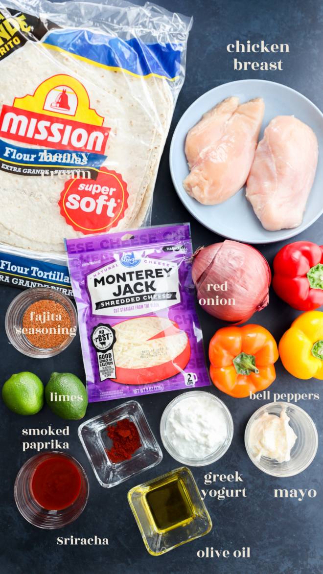 Ingredients for a chicken fajitas wrap are laid out on a surface, including chicken breasts, flour tortillas, Monterey Jack cheese, red onion, bell peppers, fajita seasoning, limes, smoked paprika, Sriracha, Greek yogurt, mayo, and olive oil.
