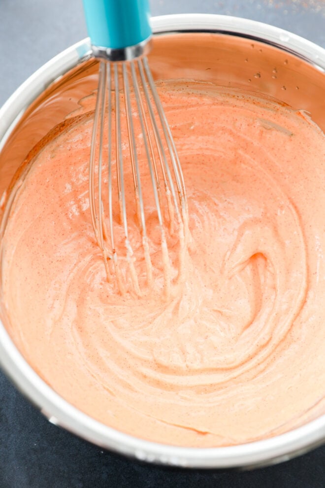 A metal whisk is stirring a creamy, sriracha yogurt mixture in a stainless steel bowl on a dark surface.