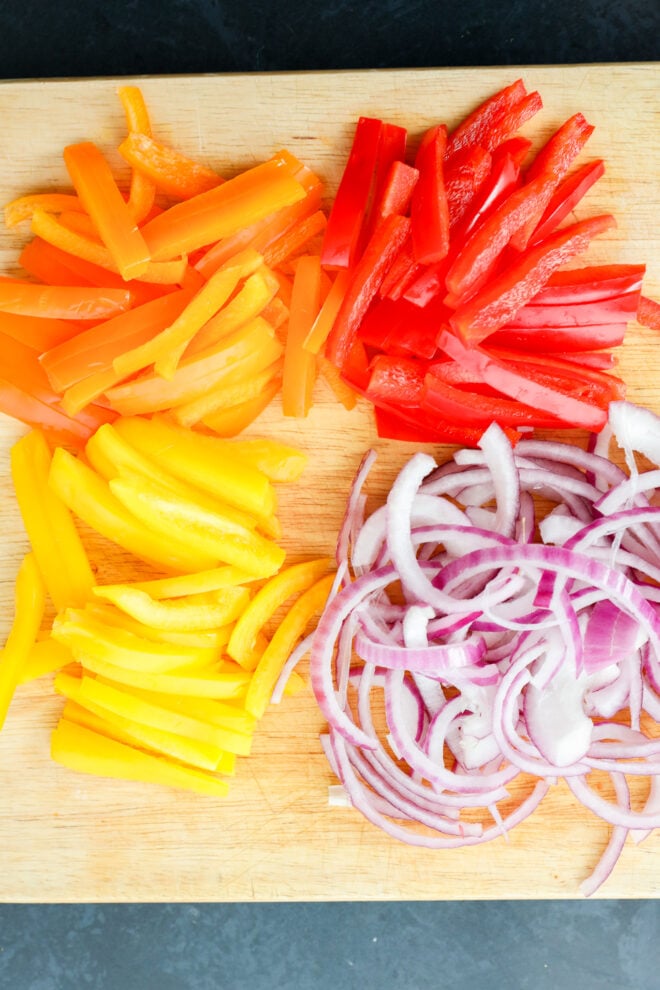 A wooden cutting board with sliced red, orange, and yellow bell peppers and thinly sliced red onion arranged in separate piles