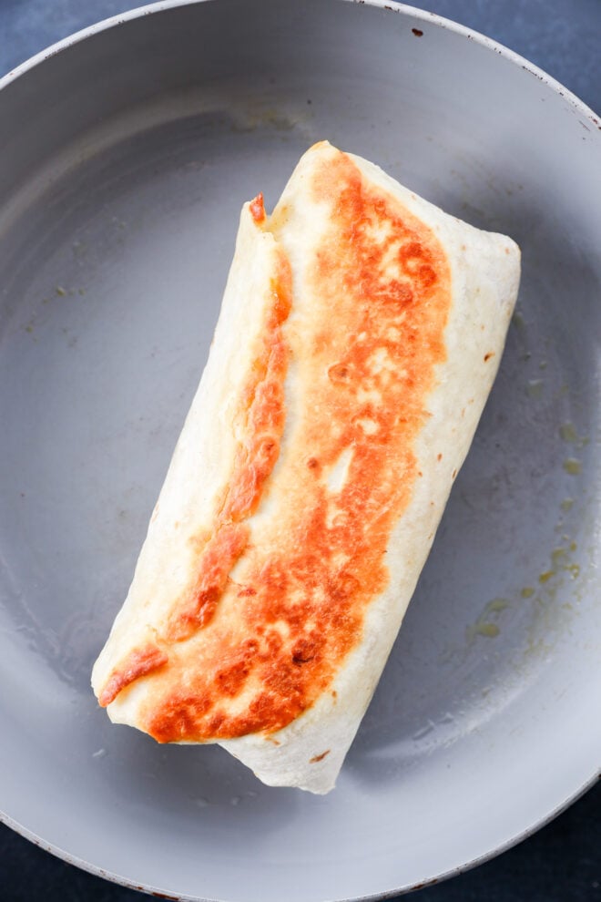 A chicken fajitas wrap with a golden, crispy side sits in a light grey skillet, showing a toasted tortilla and a slightly melted cheese edge.
