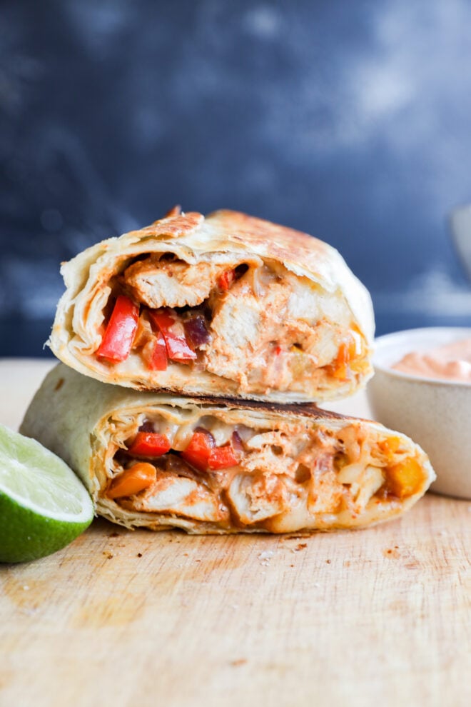 A close-up of two grilled chicken fajitas wraps stacked on a wooden surface, filled with chicken, red peppers, melted cheese, and sauce, with a lime wedge and a dip in a small bowl beside them.